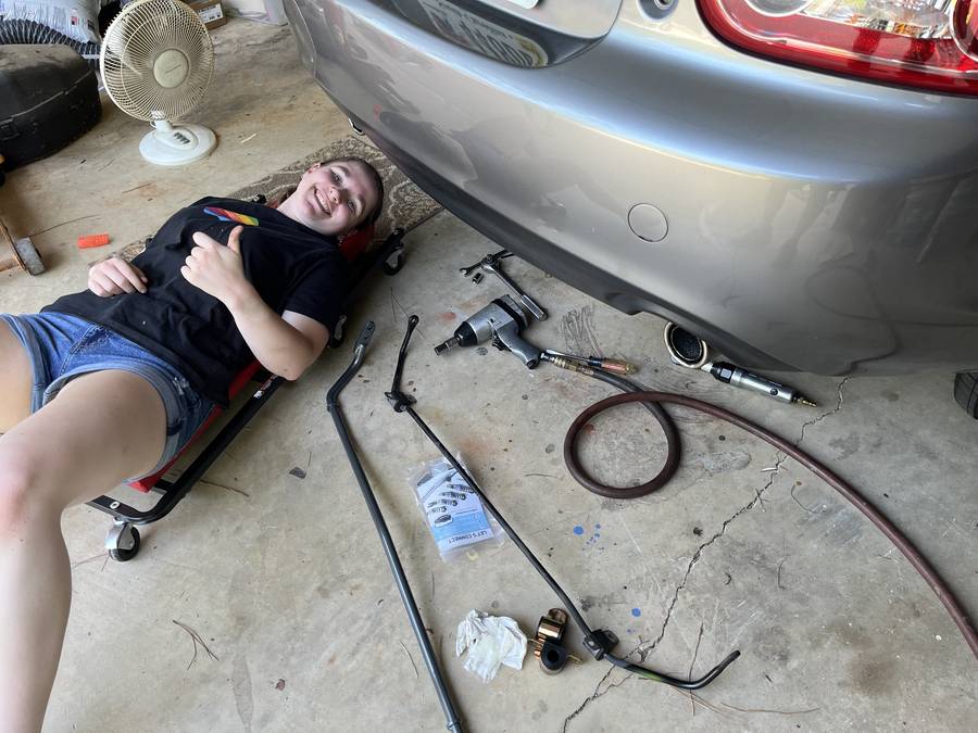 Smiling on a creeper under the rear bumper of a silver car, giving a thumbs up, surrounded by an impact wrench, sway bar, and bushings mid-replacement