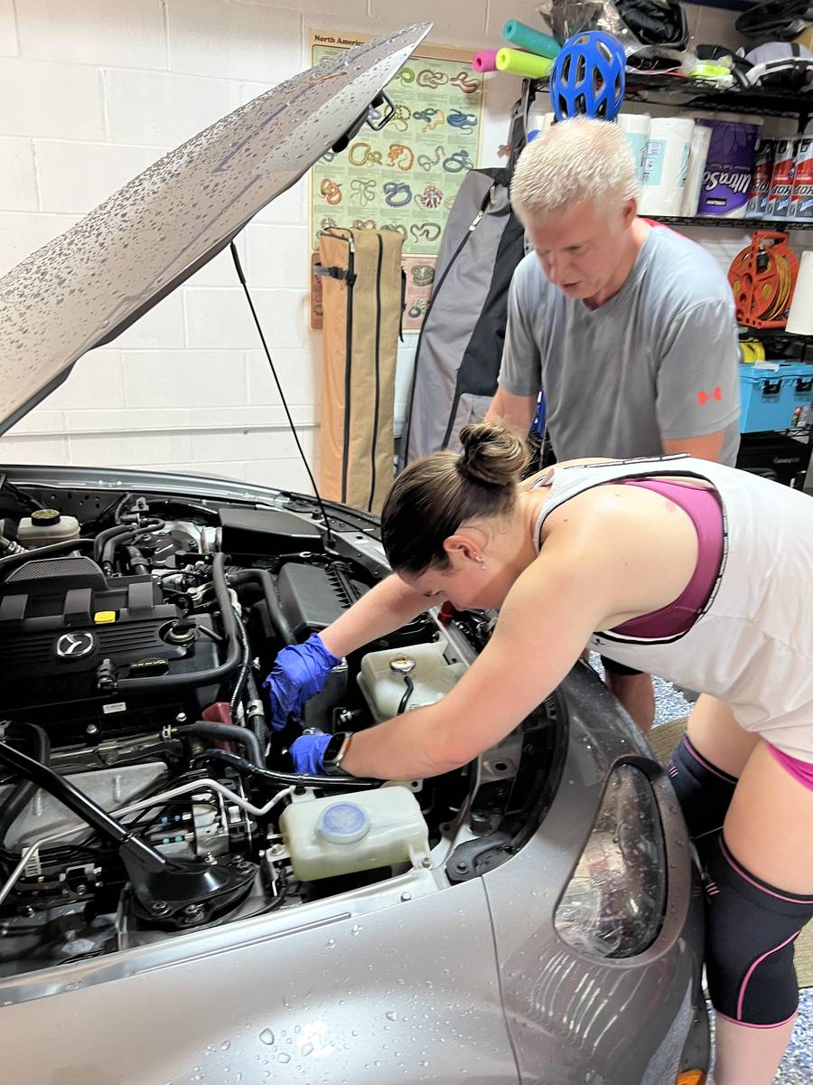 Working under the open hood on the tensioner and idler pulleys, with a second person looking on